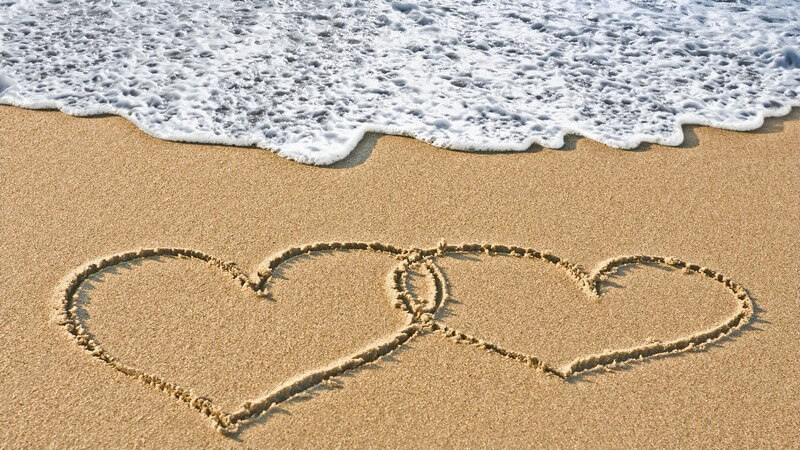 Zwei Herzen in Sand gemalt am Strand, dahinter kommt eine Welle