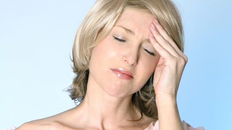 Blonde Frau mit Kopfschmerzen vor hellblauem Hintergrung fasst sich mit geschlossenen Augen an die Stirn