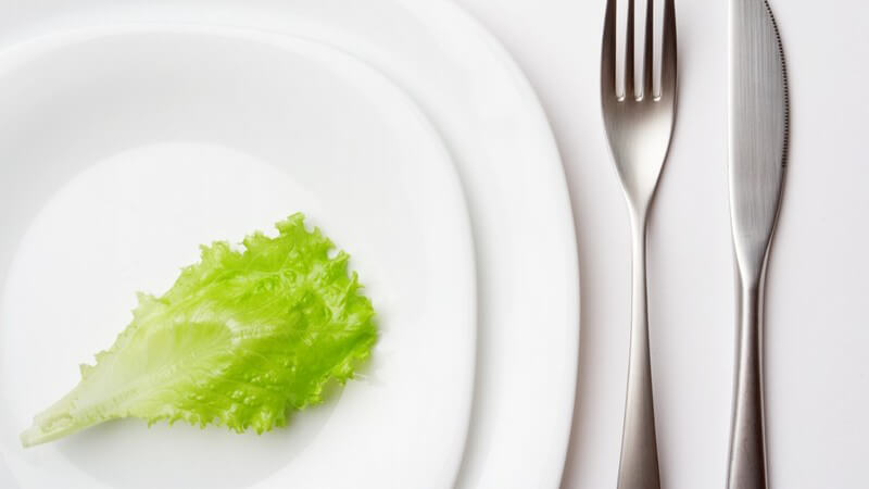 Zwei weiße Porzellanteller und Besteck mit Messer und Gabel mit einem einzelnen Salatblatt als Menü Diät Abnehmen
