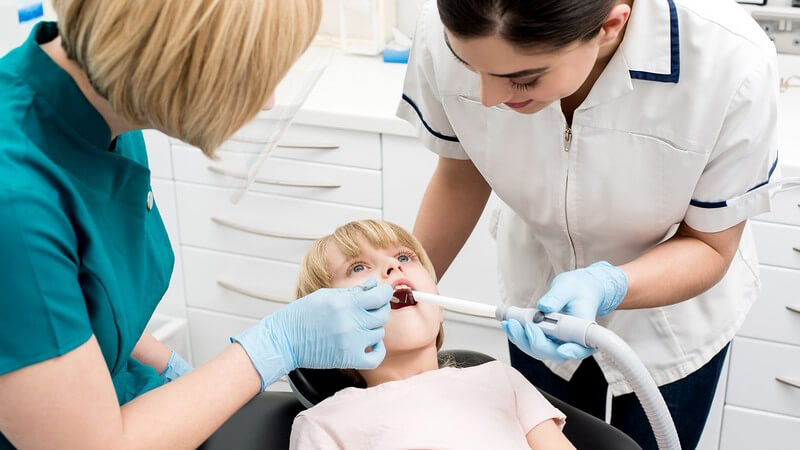 Kind liegt auf Zahnarztstuhl und wird von Zahnärztin behandelt, Zahnarzthelferin assistiert mit dem Absauger