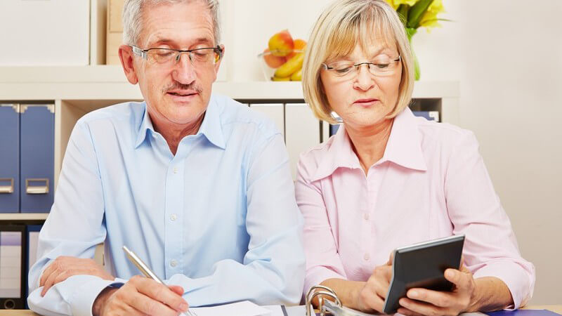 Älteres Paar sitzt am Tisch und macht die Steuererklärung, er mit Stift, sie mit Taschenrechner