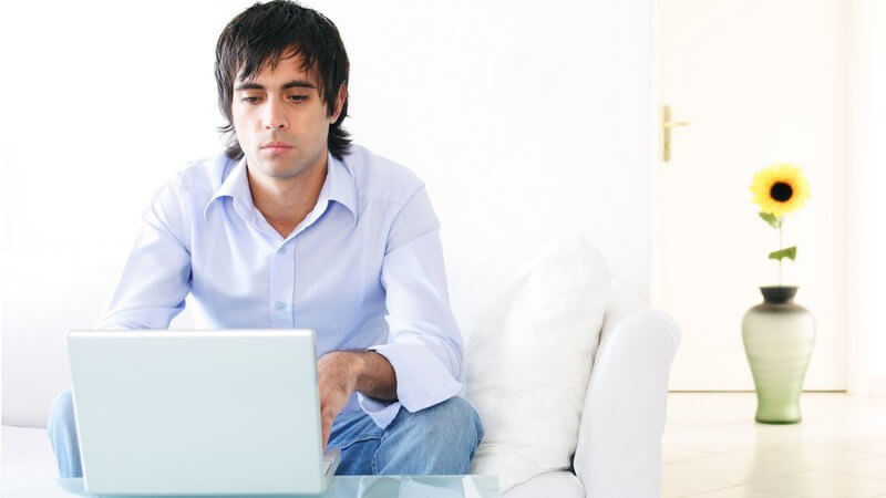Junger Mann im Hemd sitzt am Wohnzimmertisch am Notebook und arbeitet