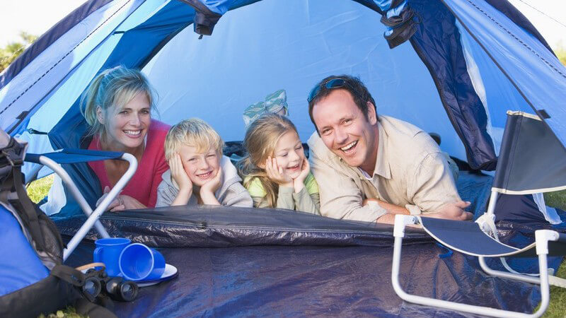 Junge Eltern liegen mit zwei kleinen Kindern im Zelt und lachen in Kamera