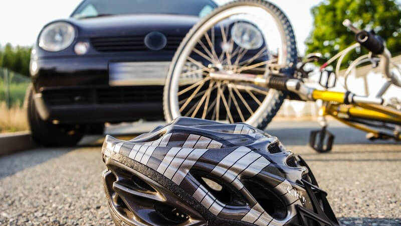 Grauer Fahrradhelm und gelbes Fahrrad liegen nach einem Unfall auf dem Asphalt vor einem Auto