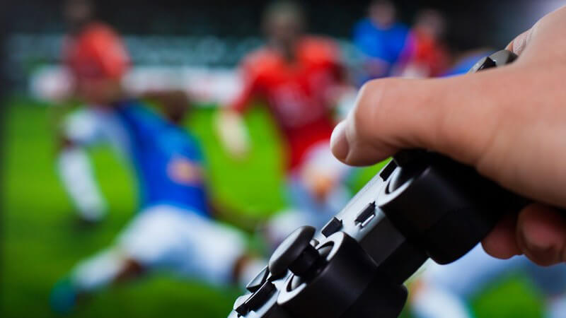 Hand mit schwarzem Gamepad vor einem Fußball-Videospiel