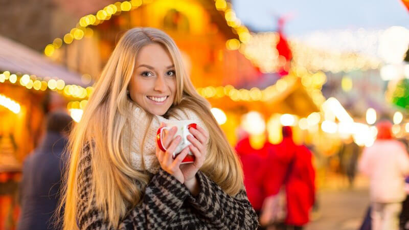 Junge Frau mit sehr langen Haaren, Schal und Wintermantel trinkt Glühwein auf dem Weihnachtsmarkt