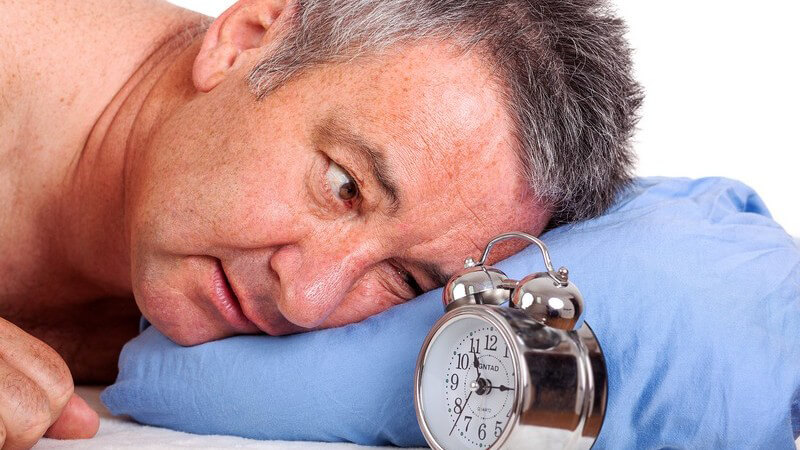 Älterer Mann liegt auf seinem blauen Kissen und schaut auf den Wecker neben ihm