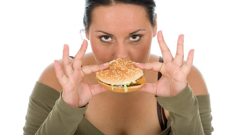 Mollige Frau hält Hamburger zwischen Daumen und Zeigefingern und schaut in Kamera