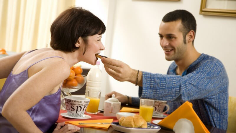Dunkelhaarige junge Frau und junger Mann, Pärchen, am Frühstückstisch, Mann füttert Frau