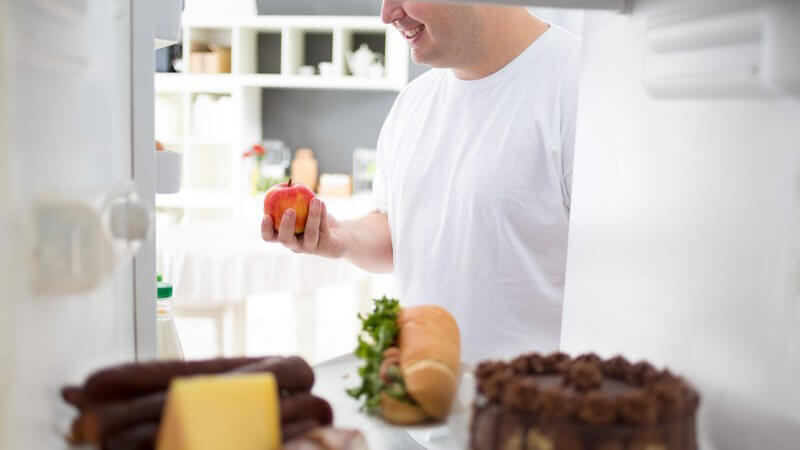 Dicker Mann steht mit Apfel in der Hand vor dem Kühlschrank, in dem sich Kuchen, Wurst und ein Baguette befinden