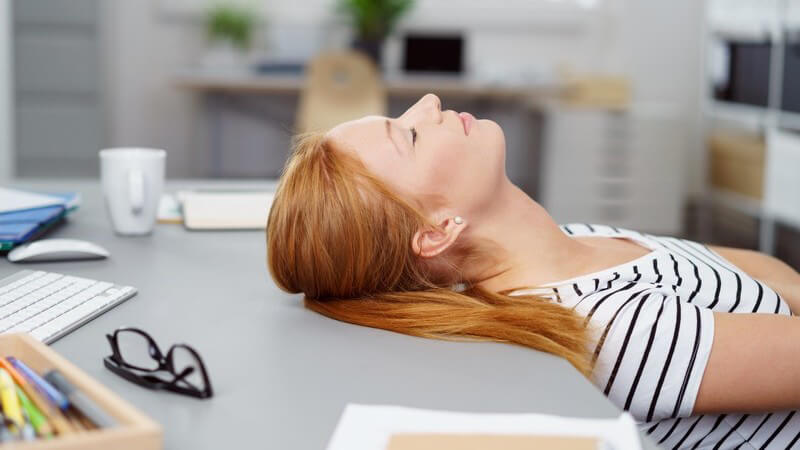Rotblonde Frau liegt mit der Kopfrückseite auf dem Schreibtisch im Büro und schläft