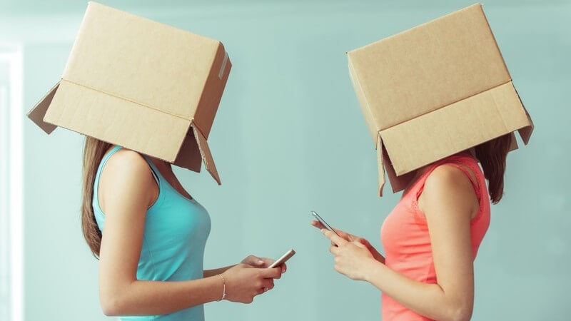 Zwei Mädchen stehen sich gegenüber, haben einen Karton über dem Kopf und schreiben sich mit dem Smartphone