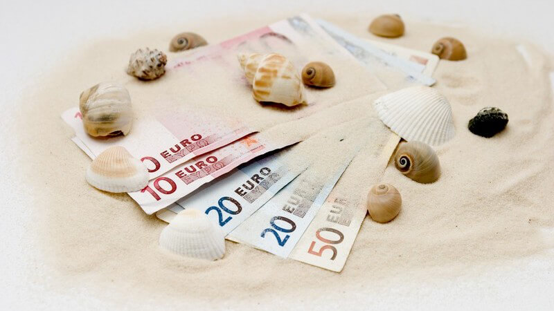 5 Geldscheine von 10-50 Euro liegen aufgefechert auf Sand mit Muscheln verziert