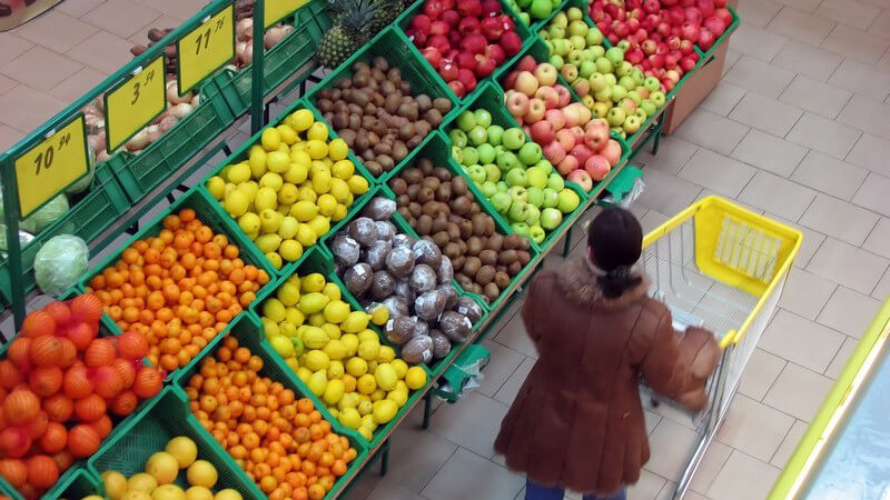 Aufnahme von oben, Frau schiebt im Supermarkt Einkaufswagen an Obstabteilung vorbei