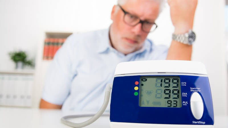 Älterer Herr mit Brille sitzt hinter einem Blutdruckmessgerät, das einen hohen Blutdruck von 199/99 anzeigt