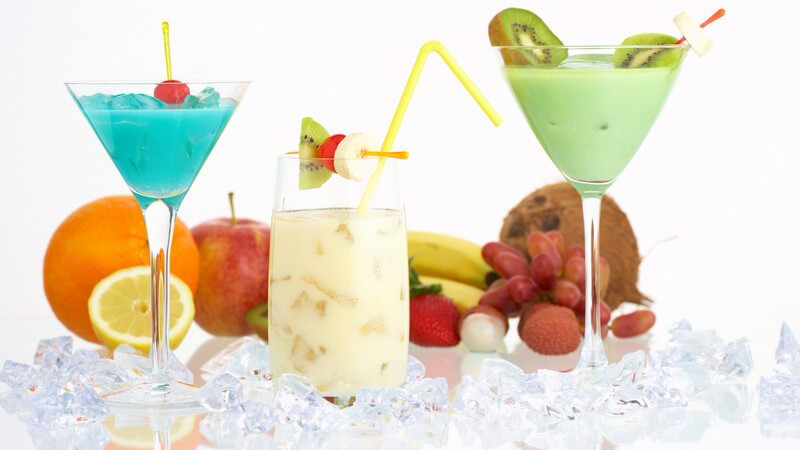 Verschiedene Cocktails in Gläsern zwischen Eiswürfeln, dahinter frisches Obst