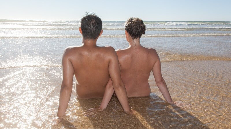Nacktes Paar sitzt am Strand im Wasser und blickt raus auf das Meer