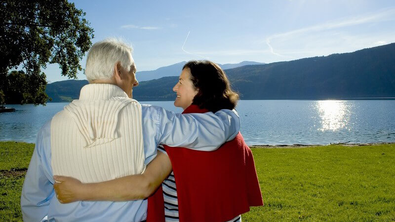 Älteres Ehepaar an Bergsee mit Sonnenschein und grüner Wiese, die Arme umeinander gelegt