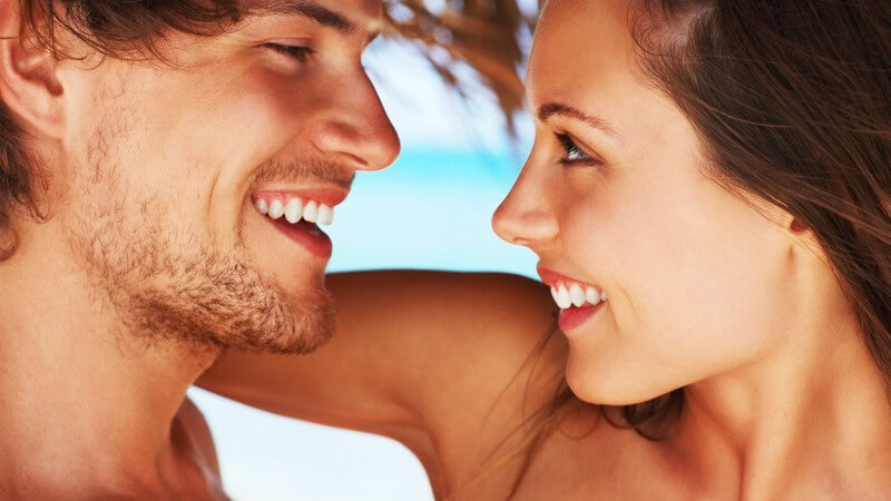 Junges Paar am Strand, beide lachen, schauen sich in die Augen