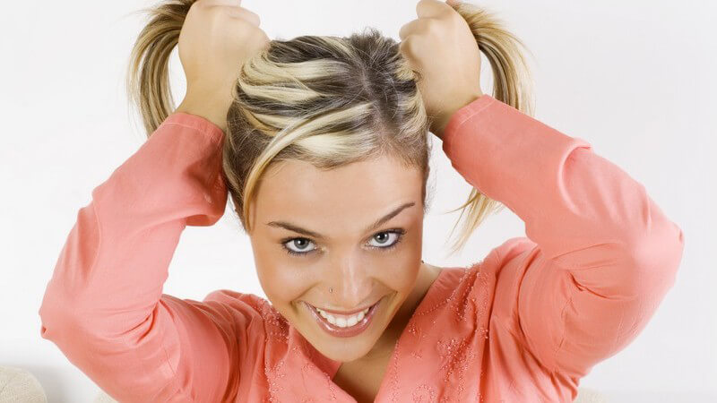 Blondierte junge Frau hält sich die Haare zu zwei Zöpfen, grinst, in lachsfarbenem Oberteil