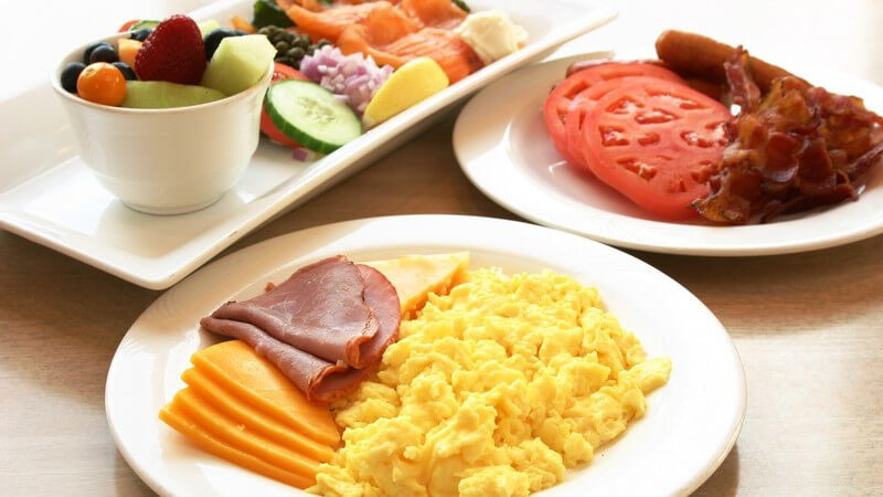 Üppiges Frühstück mit zwei Porzellantellern mit Rührei, Käse, Roastbeef, Tomaten, Lachs, Früchten und Gemüse