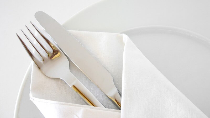 Sitzplatz mit Besteck (Messer und Gabel) in Serviette auf weißem Teller vor weißem Hintergrund, edel und schlicht