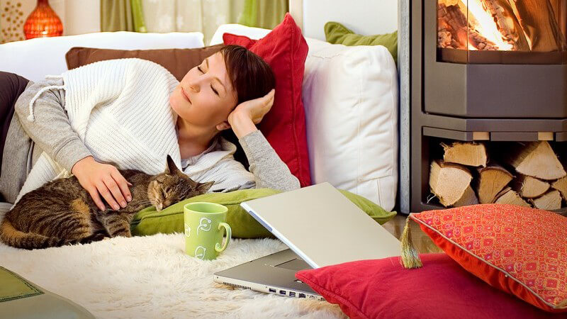 Junge Frau liegt mit Katze auf Couch vor Kamin