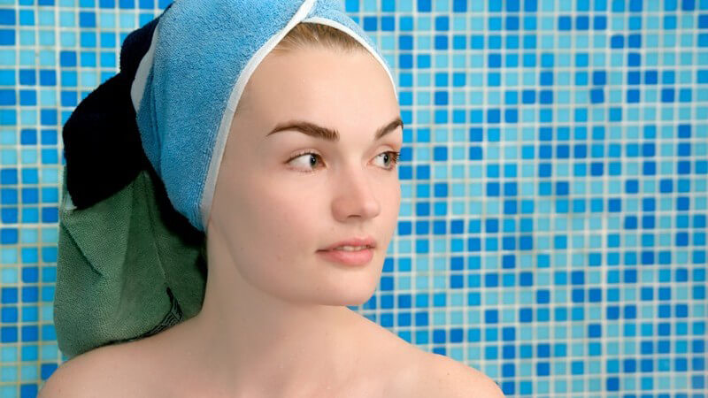 Frau mit Handtuch auf Kopf, im Hintergrund blau-türkise Badezimmer Kacheln