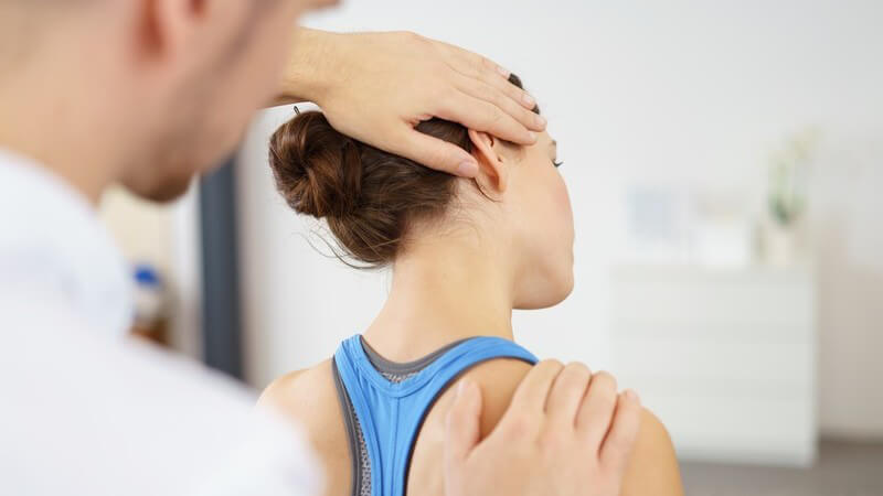 Junger Physiotherapeut behandelt eine junge Patientin mit Dutt wegen Nackenschmerzen