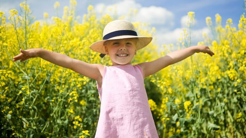 Kleines Mädchen mit Sonnenhut steht lachend mit Armen ausgestreckt vor gelbem Blumenfeld
