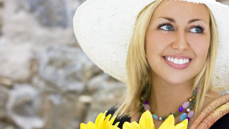 Blonde Frau mit weißem Strohhut lächelt, hält Sonnenblumen in Hand