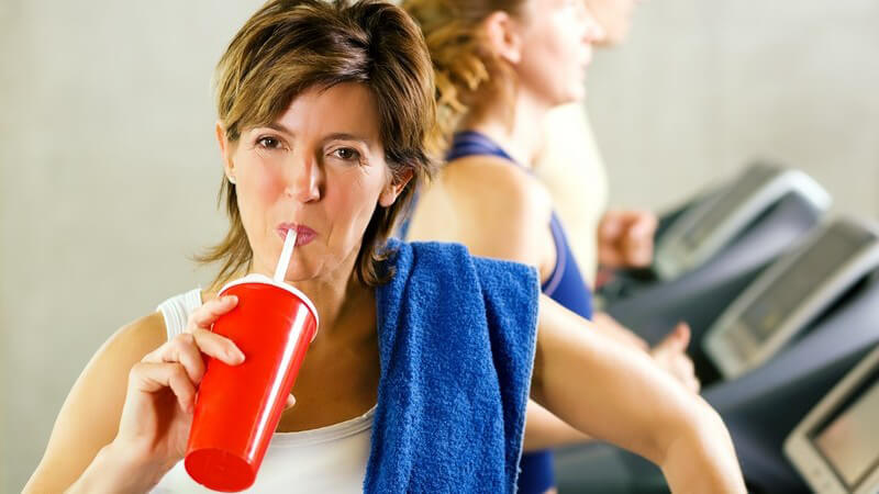 Frau auf Laufband in Fitnessstudio mit blauem Handtuch auf der Schulter trinkt auf Trinkbecher, Sportler im Hintergrund