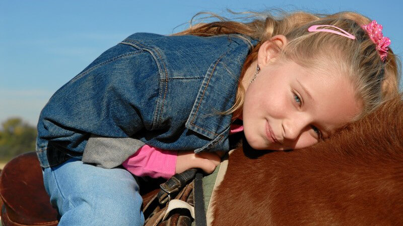Blondes Mädchen in Jeansjacke sitzt auf braunem Pferd, den Kopf auf dessen Rücken gelegt