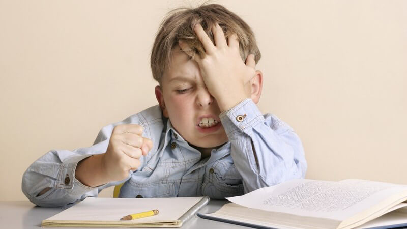Junge in hellblauem Hemd sitzt mit geballter Faust frustriert und wütend über seinen Hausaufgaben