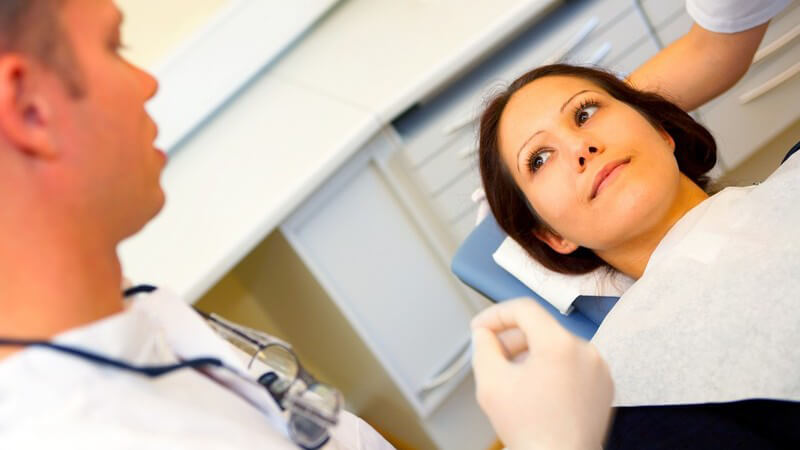 Brünette Frau auf Zahnarztstuhl, links verschwommen der Arzt mit Brille um den Hals