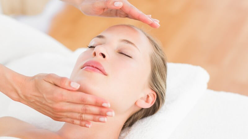 Frau liegt mit geschlossenen Augen entspannt bei einer Reiki-Behandlung auf dem Rücken, Hände schweben über ihrem Kopf