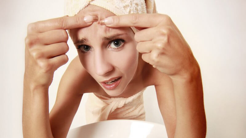 Junge Frau in Handtuch steht vor Spiegel, drückt Pickel auf Stirn aus