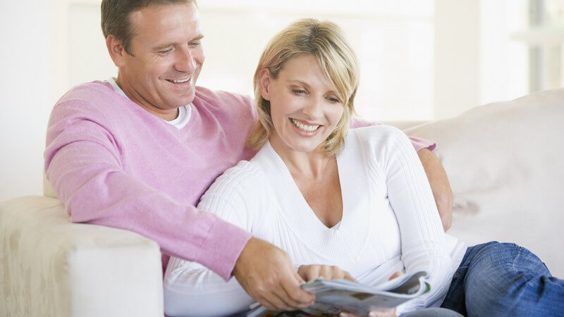 Erwachsenes Paar sitzt lächelnd auf der Couch und blättert in einer Zeitschrift