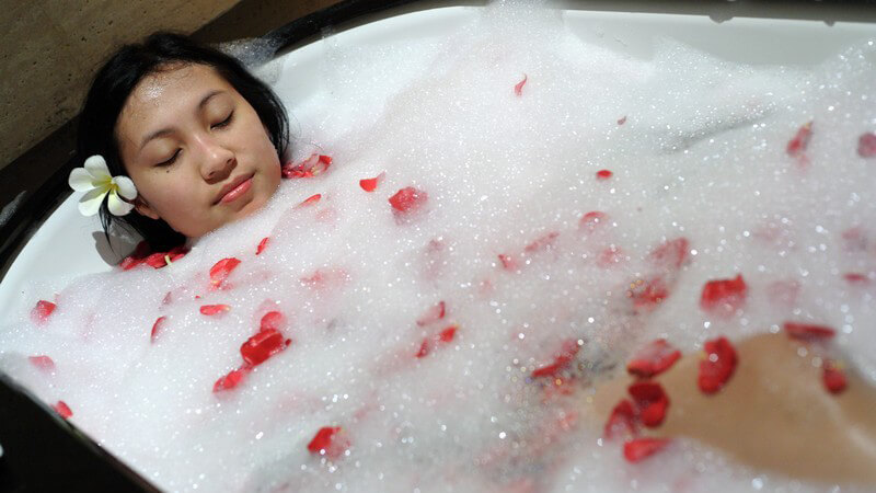 Dunkelhaarige junge Frau mit geschlossenen Augen in Badewanne mit viel Schaum und Rosenblättern