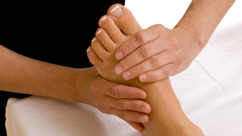 Linker manikürierter Fuß einer Frau liegt auf weißem Laken und wird von zwei Händen einer Frau massiert (Fussmassage)