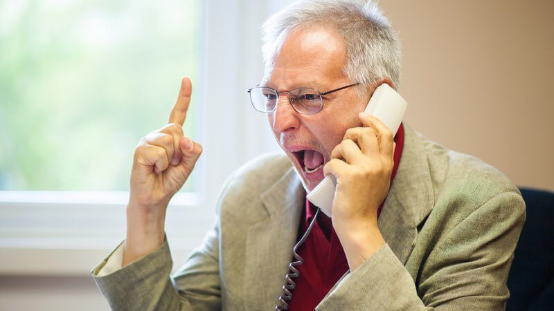 Mann in hellgrauem Sacko und mit Brille sitzt am Telefon und schreit hinein, erhobener Zeigefinger