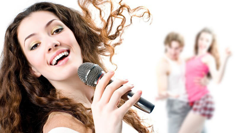 Junge, dunkelhaarige Frau singt mit Mikrofon, im Hintergrund ein tanzendes Paar