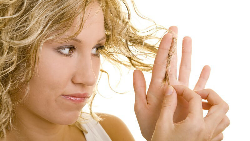 Junge blonde Frau hält Haarspitzen in Hand, schaut kritisch