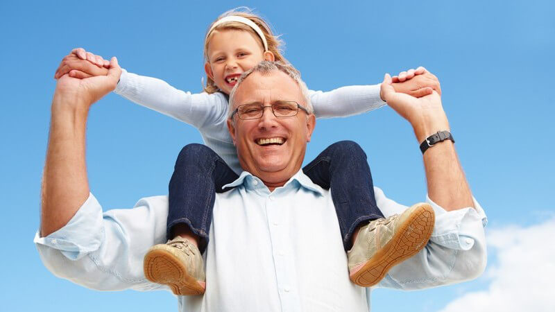 Großvater trägt seine Enkelin auf Schulter unter blauem Himmel, beide lachen