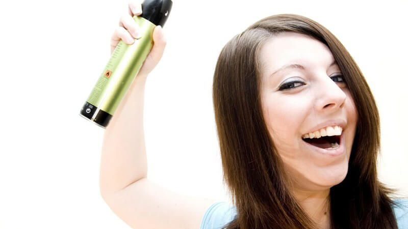 Braunhaarige Frau sprüht sich Haarspray auf Haare, lacht in Kamera