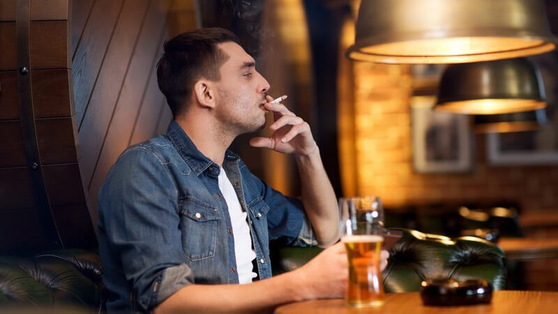 Mann in Jeanshemd sitzt am Tisch in einer Kneipe, trinkt Bier und raucht Zigarette
