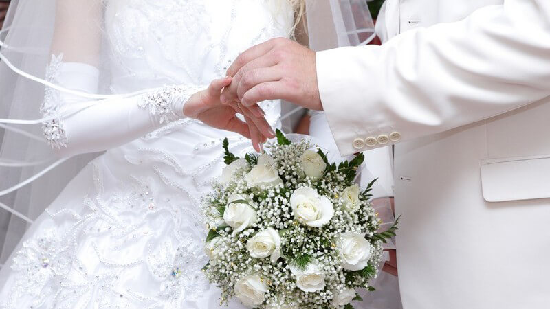 Nahaufnahme Brautpaar in weiß, Bräutigam zieht Braut den Ehering an