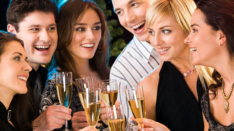 Sechs lachende Freunde in festlicher Kleidung stoßen mit Glas Sekt an
