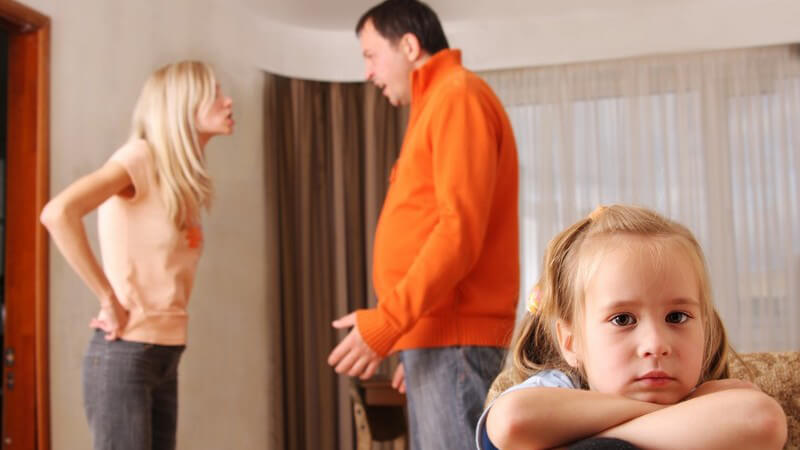 Kleines Mädchen schaut traurig in Kamera, im Hintergrund streiten sich Eltern