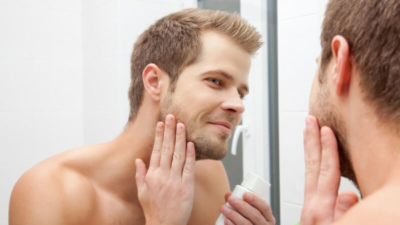 Gepflegter Mann mit leichtem Vollbart steht vor dem Spiegel und reibt seine Wange mit Aftershave ein